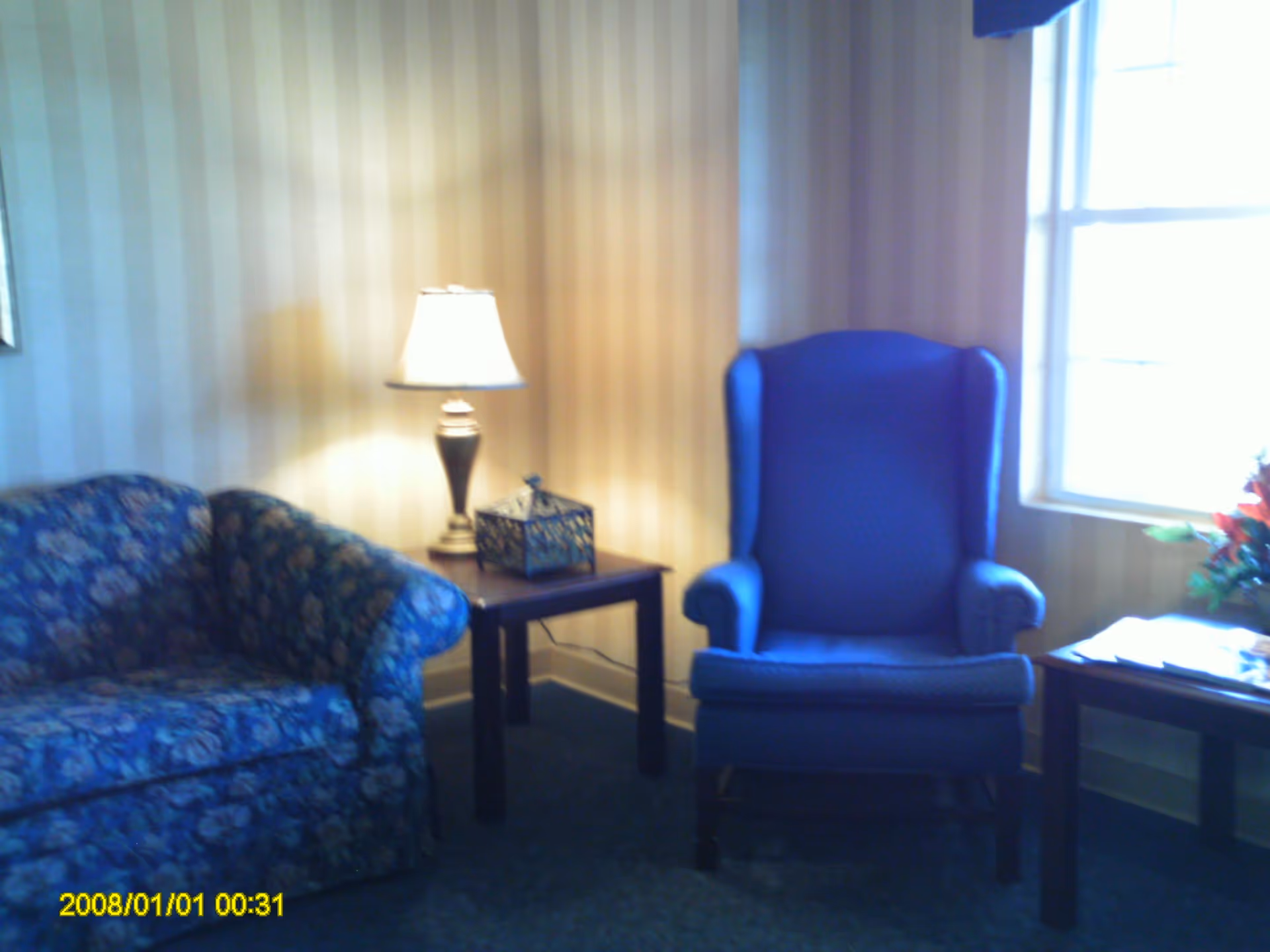 Small sitting area with a floral couch, a blue wingback chair, side tables and a lamp by a window.