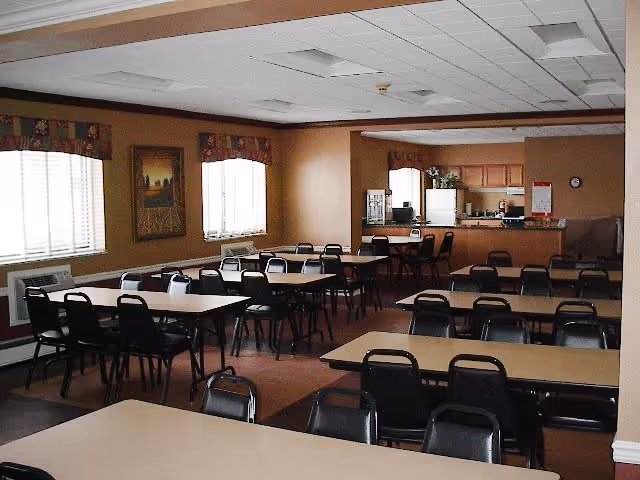 Communal dining room with rows of tables and chairs and a serving kitchen at the back.
