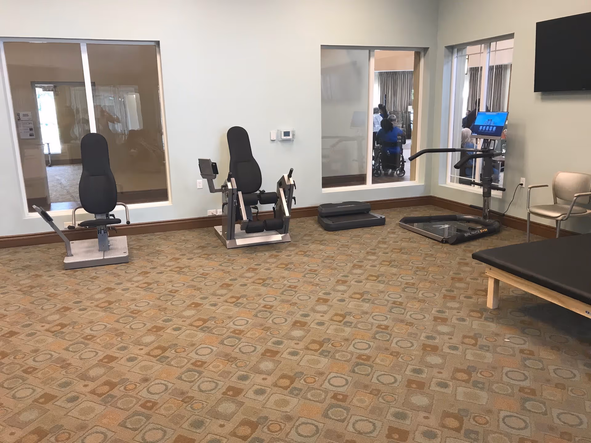 Exercise/physical therapy room with seated exercise machines, a treadmill, a treatment table, and windows into adjacent areas.
