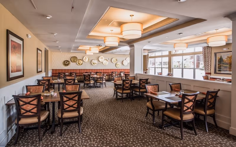 A spacious dining room with multiple wooden tables and cushioned chairs arranged neatly. The room features patterned carpet flooring, large windows allowing natural light, decorative wall art including framed pictures and circular metallic plates, and ceiling lights with drum-shaped shades.