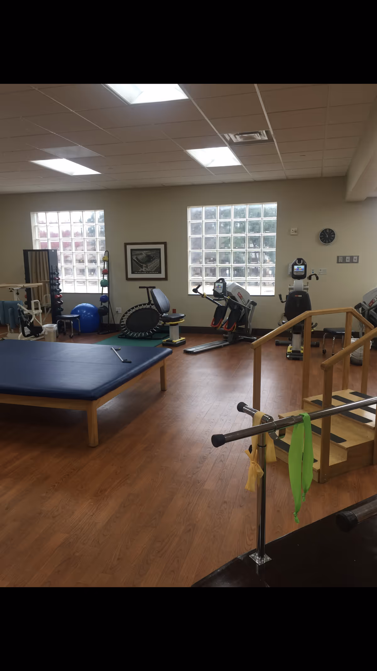 A rehabilitation and exercise room with wooden flooring, featuring exercise equipment such as stationary bikes, a trampoline, parallel bars with resistance bands, a padded therapy table, and a set of stairs with handrails for physical therapy. The room has large glass block windows allowing natural light to enter.