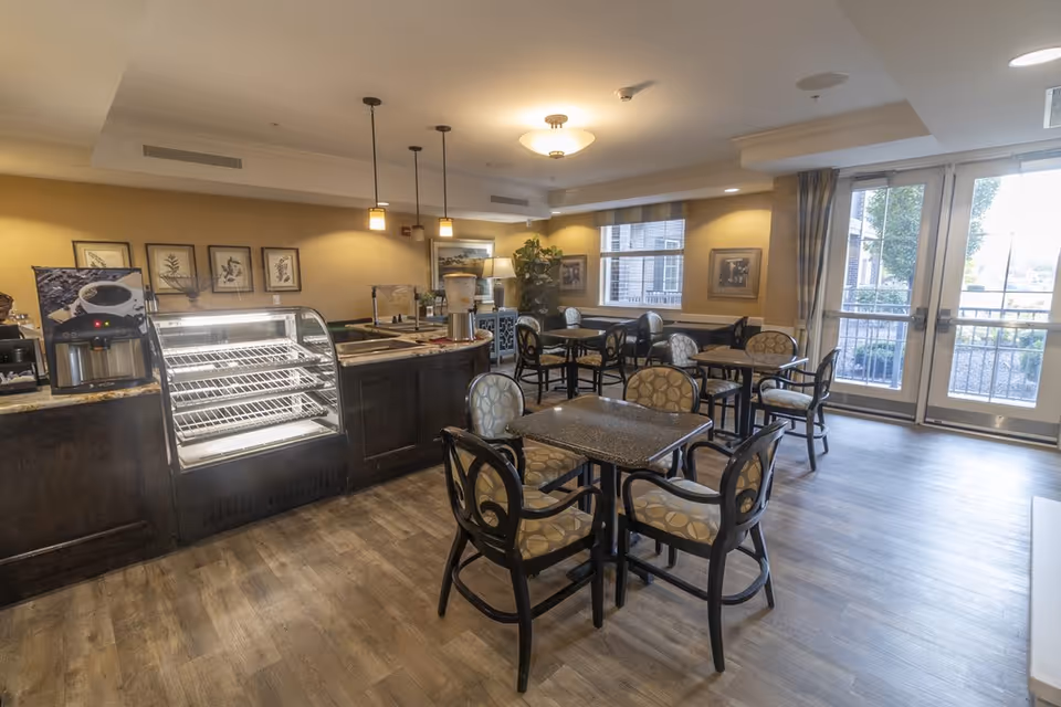 A well-lit dining area in Independence Village of Carmel featuring several tables with cushioned chairs arranged neatly. There is a counter with a glass display case and a coffee machine on the left side. Large windows and glass doors allow natural light to enter the room, and framed artwork decorates the walls.