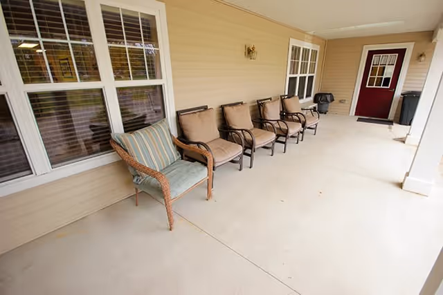 Covered front porch with several cushioned chairs lined up against beige siding and a red door at the far end.