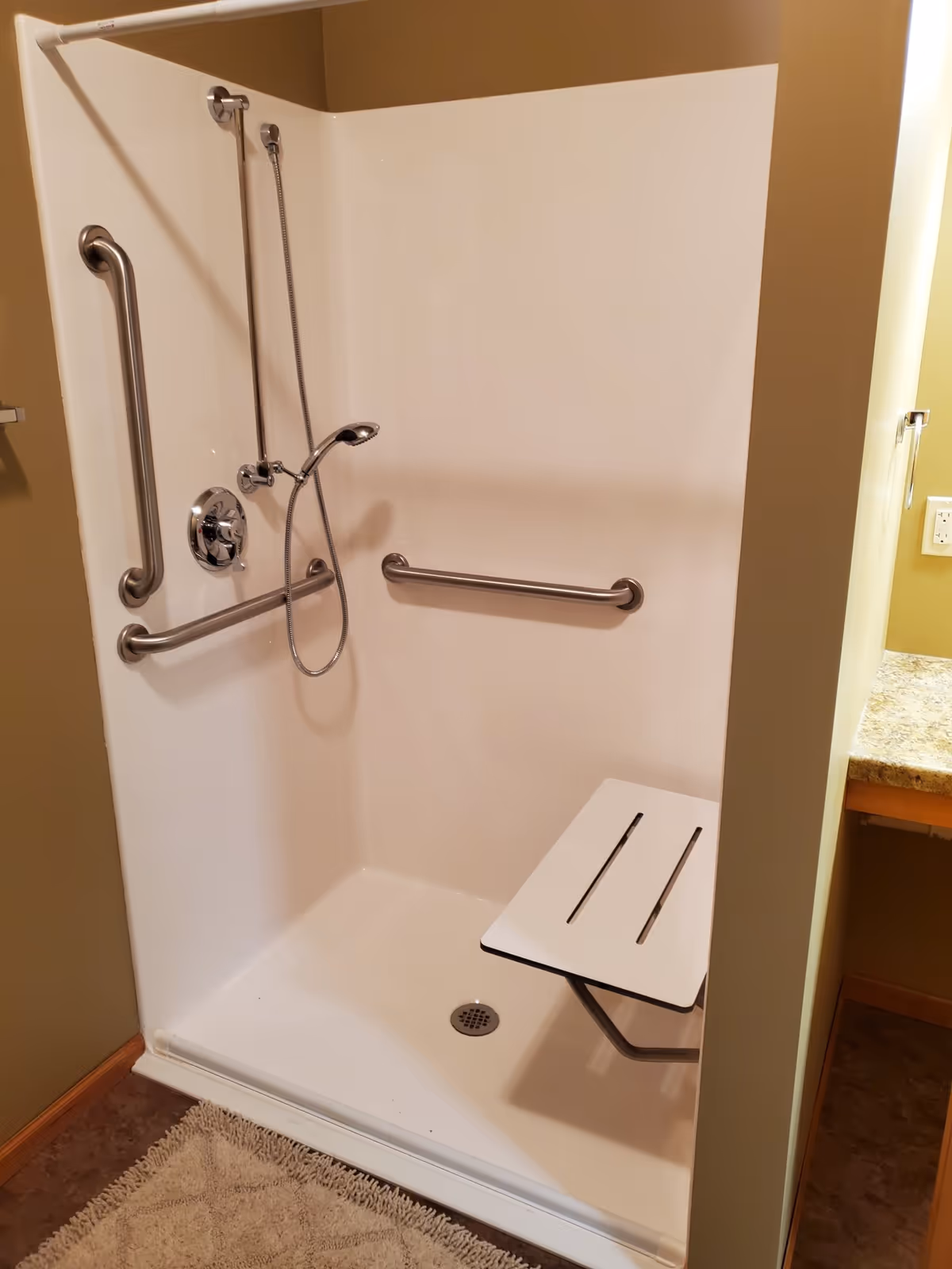 A walk-in shower with white walls, equipped with multiple stainless steel grab bars and a handheld showerhead. There is a foldable white shower seat attached to the wall. The shower floor is white with a drain in the center. A beige bath mat is placed outside the shower on the floor.