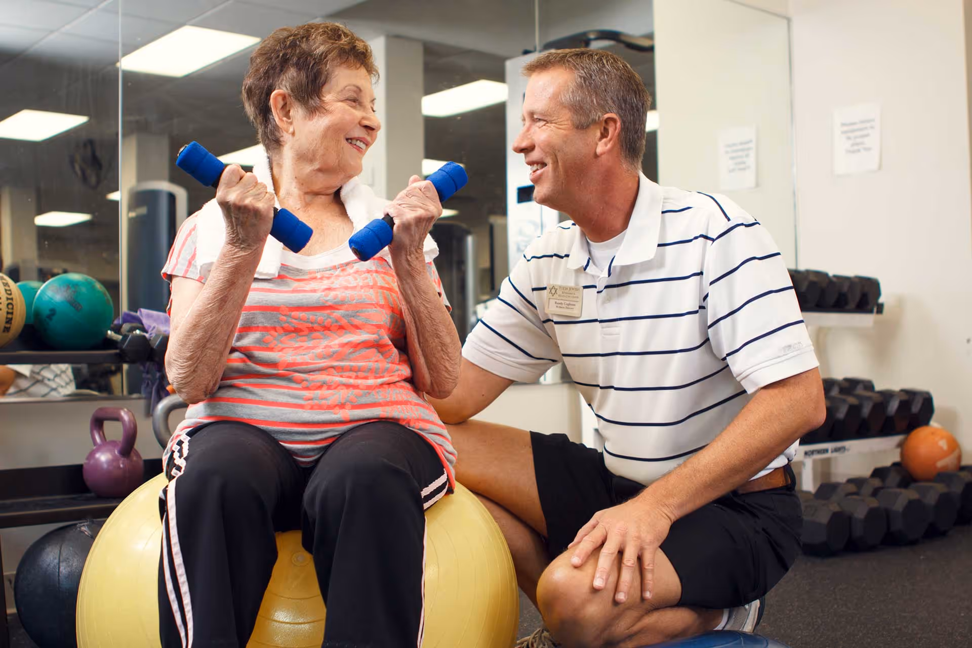An elderly woman sitting on a yellow exercise ball holding blue dumbbells, smiling and looking at a man kneeling beside her who is wearing a striped polo shirt and black shorts in a gym setting with weights and exercise equipment in the background.