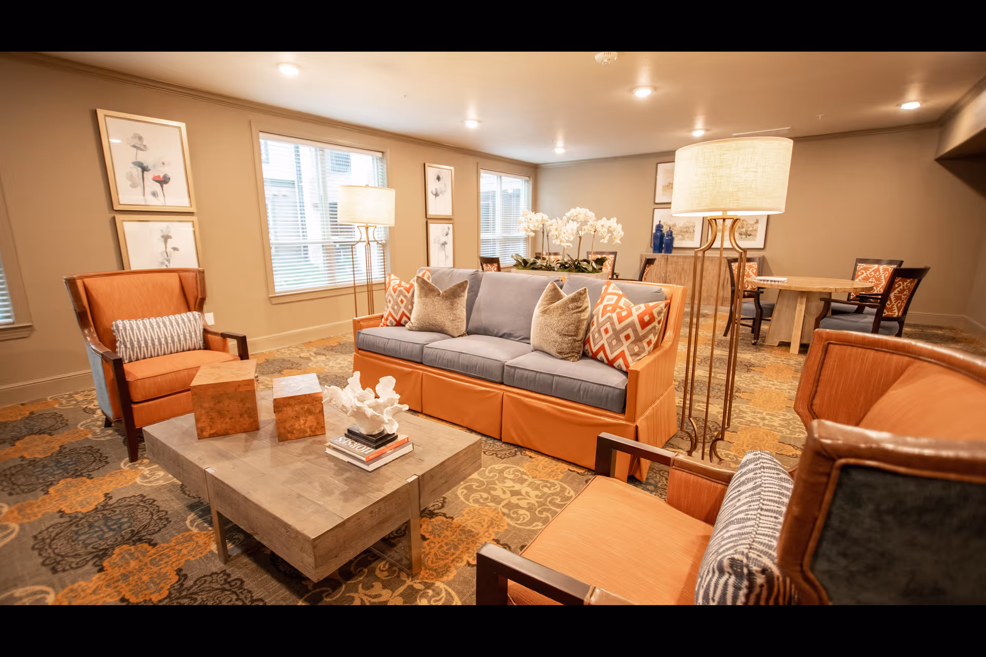 A cozy living room area in an assisted living facility featuring a blue and orange sofa with patterned pillows, two orange armchairs with cushions, a wooden coffee table with decorative items, a floor lamp, and framed floral artwork on the walls. The room has large windows with blinds and a patterned carpet.