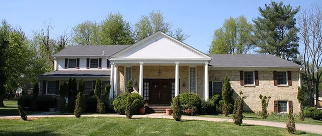Front exterior view of a large two-story residential building with a white portico supported by columns, surrounded by green trees and shrubs, under a clear blue sky.