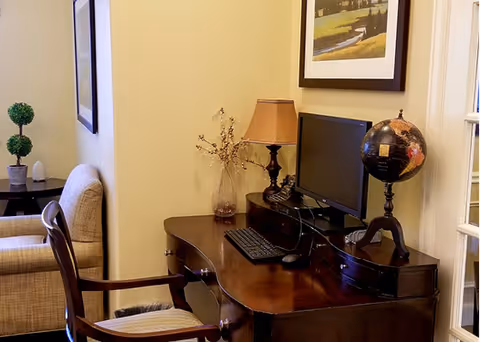 A cozy interior corner featuring a dark wooden desk with a computer monitor, keyboard, mouse, a decorative globe, and a table lamp. Next to the desk is a wooden chair with a cushioned seat. In the background, there is a beige upholstered armchair and a small side table with a potted plant. A framed picture hangs on the wall above the desk.