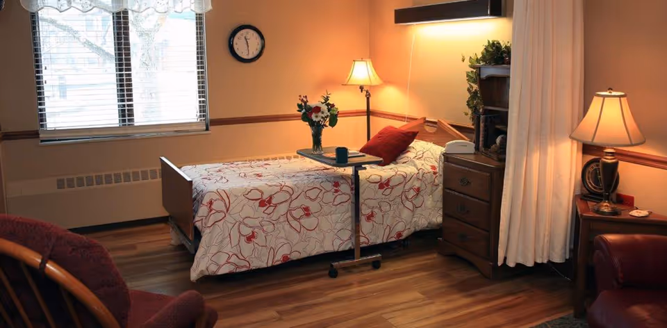 A cozy bedroom in a senior living facility with a single bed covered in a floral patterned bedspread. There is a small overbed table with a vase of flowers and a cup on it. The room has wooden flooring, a window with blinds, a clock on the wall, a floor lamp, a bedside table with a lamp, and a curtain partially drawn to one side.