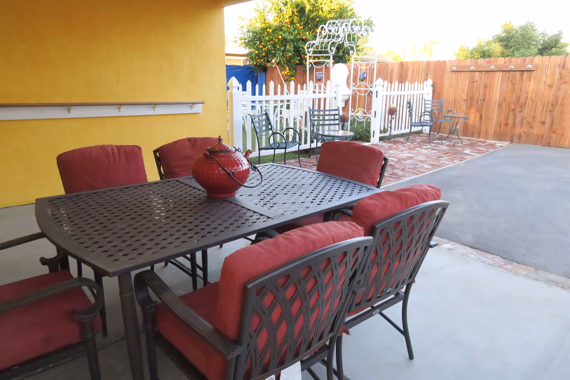 Covered outdoor patio with a metal dining table and red-cushioned chairs, yellow wall, white picket fence and a brick patio beyond.