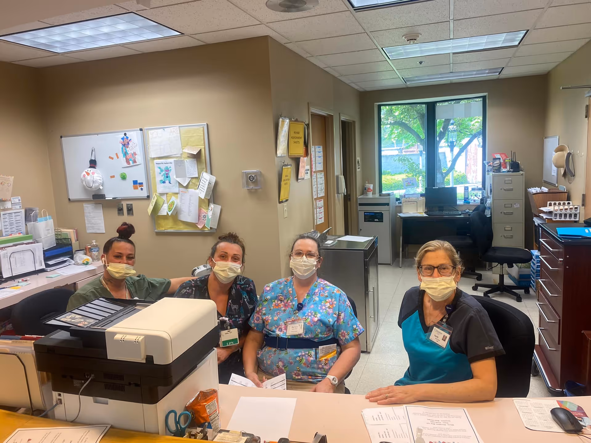 Four healthcare workers wearing face masks sitting behind a reception desk in an office area with computers, papers, and office supplies. The room has beige walls, a window showing greenery outside, and bulletin boards with pinned papers.