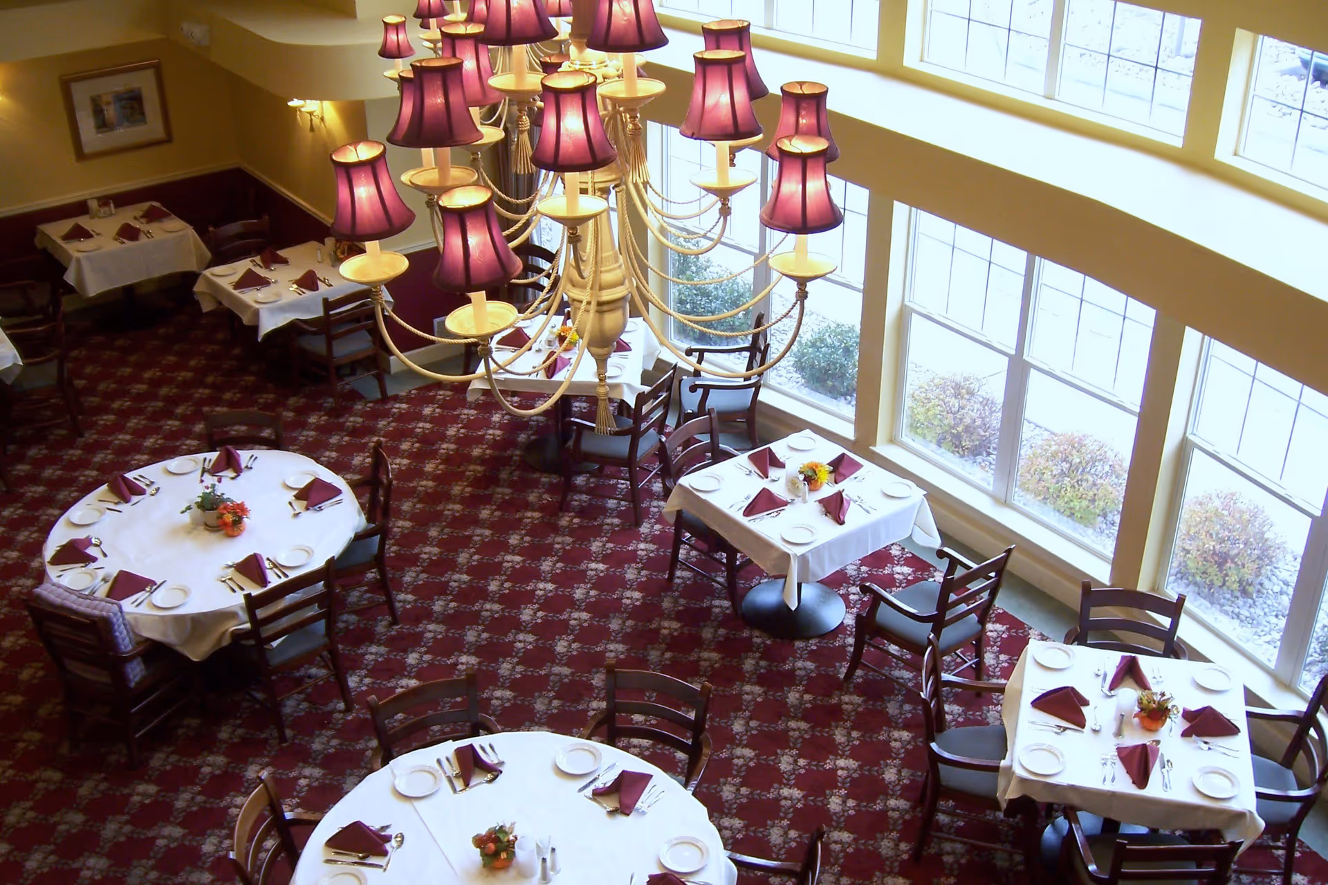 Overhead view of a dining room with round and square tables covered with white tablecloths, each set with plates, silverware, and folded burgundy napkins. A large chandelier with multiple purple lampshades hangs above the tables. Large windows let in natural light and show some shrubbery outside.