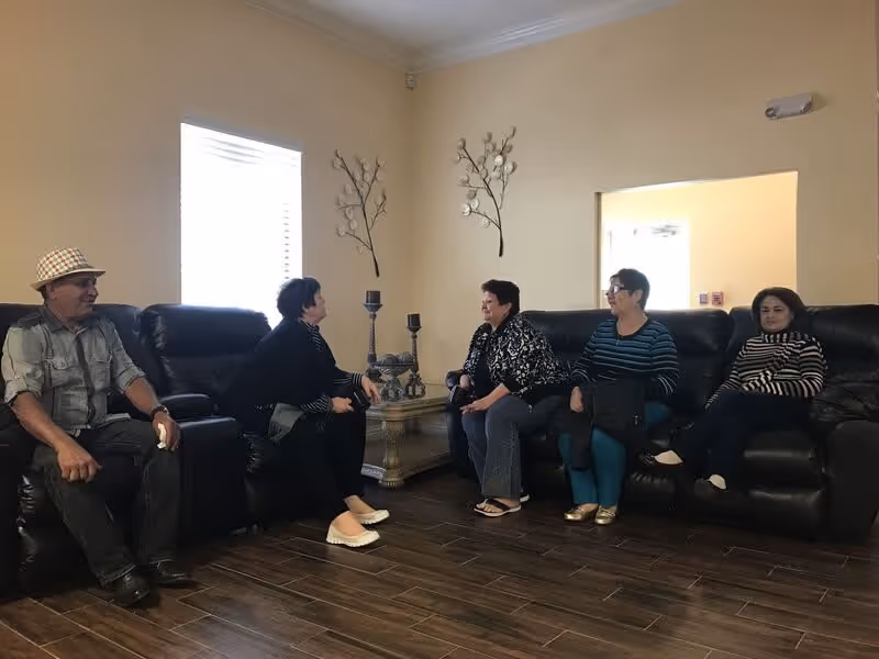 Five adults sitting and chatting in a living room with dark leather sofas, a wooden coffee table with decorative candle holders, and wall decorations shaped like branches with flowers. The room has a window with blinds and light-colored walls.