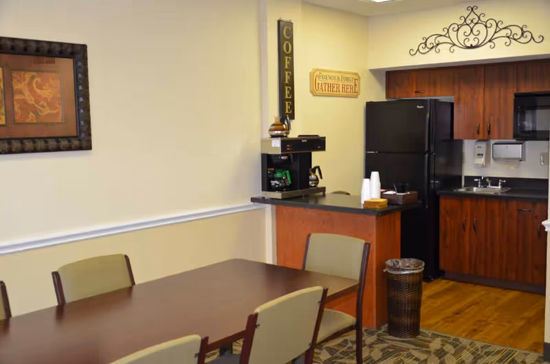 A small kitchen and dining area with a wooden table and four chairs. The kitchen has wooden cabinets, a black refrigerator, a coffee maker on the counter, a sink, and a trash bin. Wall decorations include a vertical sign that says 'COFFEE' and a sign that reads 'Friends & Family Gather Here.'