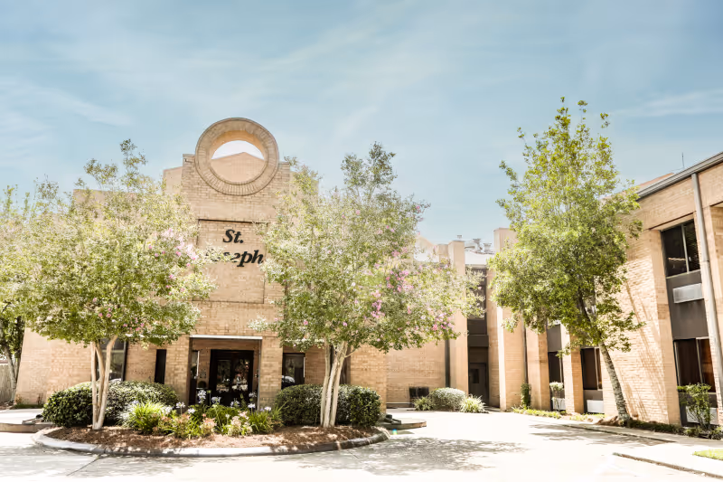 Exterior front view of St. Joseph Nursing Home building with light brown brick walls, a circular architectural feature above the entrance, and trees with green foliage and pink flowers in front of the building under a blue sky.