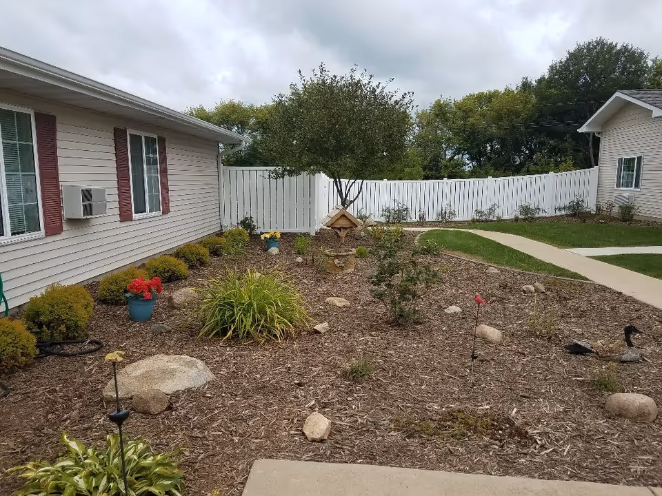 Outdoor garden area with mulch, small bushes, flowers in pots, decorative birdhouse, and a white fence in the background. There are two buildings visible on either side with beige siding and red shutters. A concrete pathway curves through the grassy area.