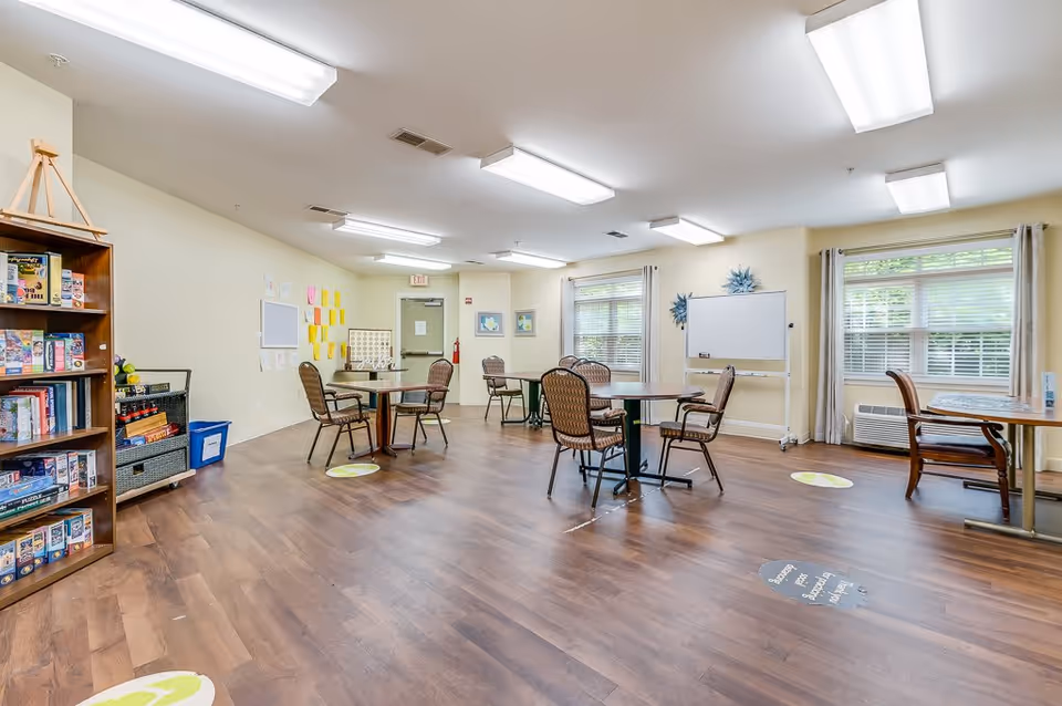 A spacious activity room with several round tables and chairs arranged for group activities. The room has large windows with curtains, a whiteboard on wheels, and a bookshelf filled with board games and puzzles. The floor is wooden, and there are social distancing floor markers. The walls are light-colored, and the room is brightly lit with ceiling lights.