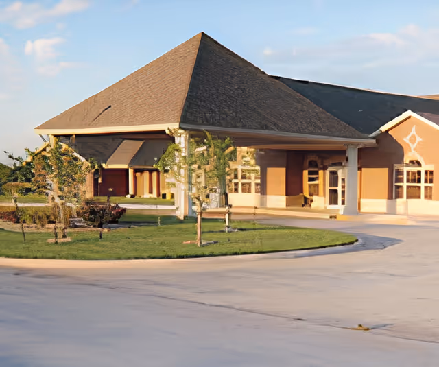 Front entrance of a senior living building with a large covered porte-cochère, arched windows, and a circular driveway with small landscaped islands.