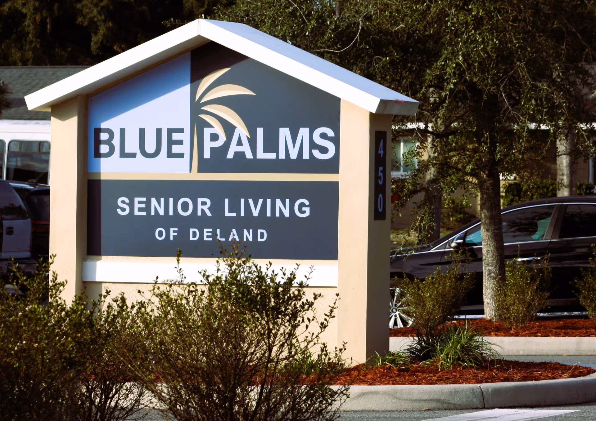 Outdoor sign for Blue Palms Senior Living of Deland with a palm tree graphic, surrounded by bushes and parked cars in the background.