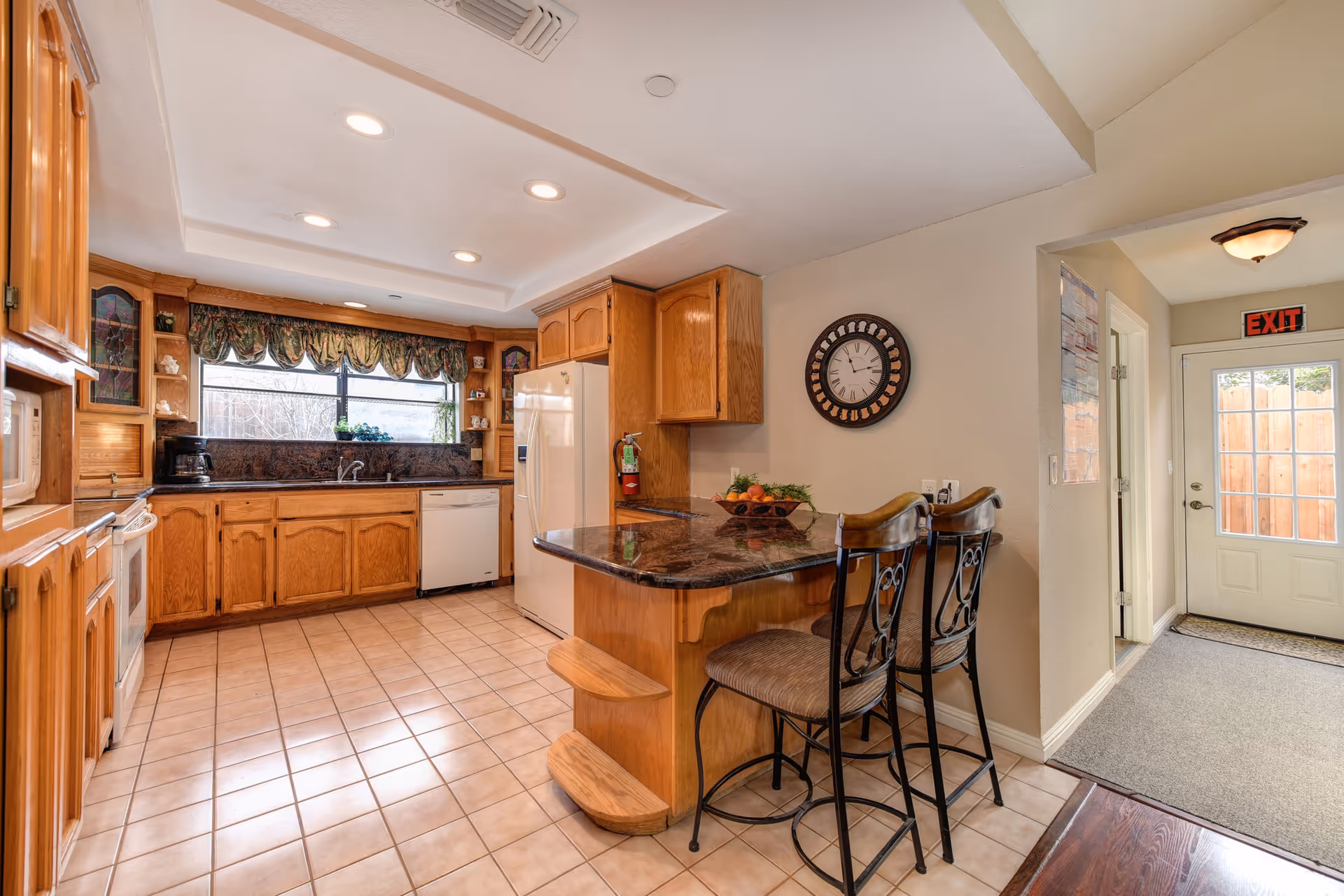 Spacious kitchen with oak cabinets, tiled floor, granite-topped island with two bar stools and a door showing an EXIT sign.