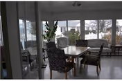 Sunlit dining/sitting area with a table and upholstered chairs surrounded by large windows overlooking a snowy yard.