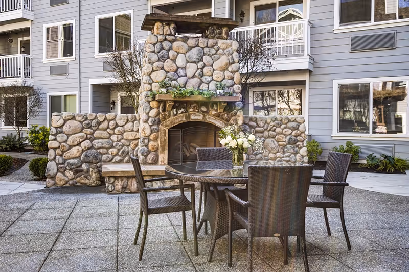 Outdoor patio area with a round glass table surrounded by four wicker chairs. In the background, there is a large stone fireplace with a wooden mantel decorated with greenery. The patio is part of a multi-story residential building with balconies and windows.