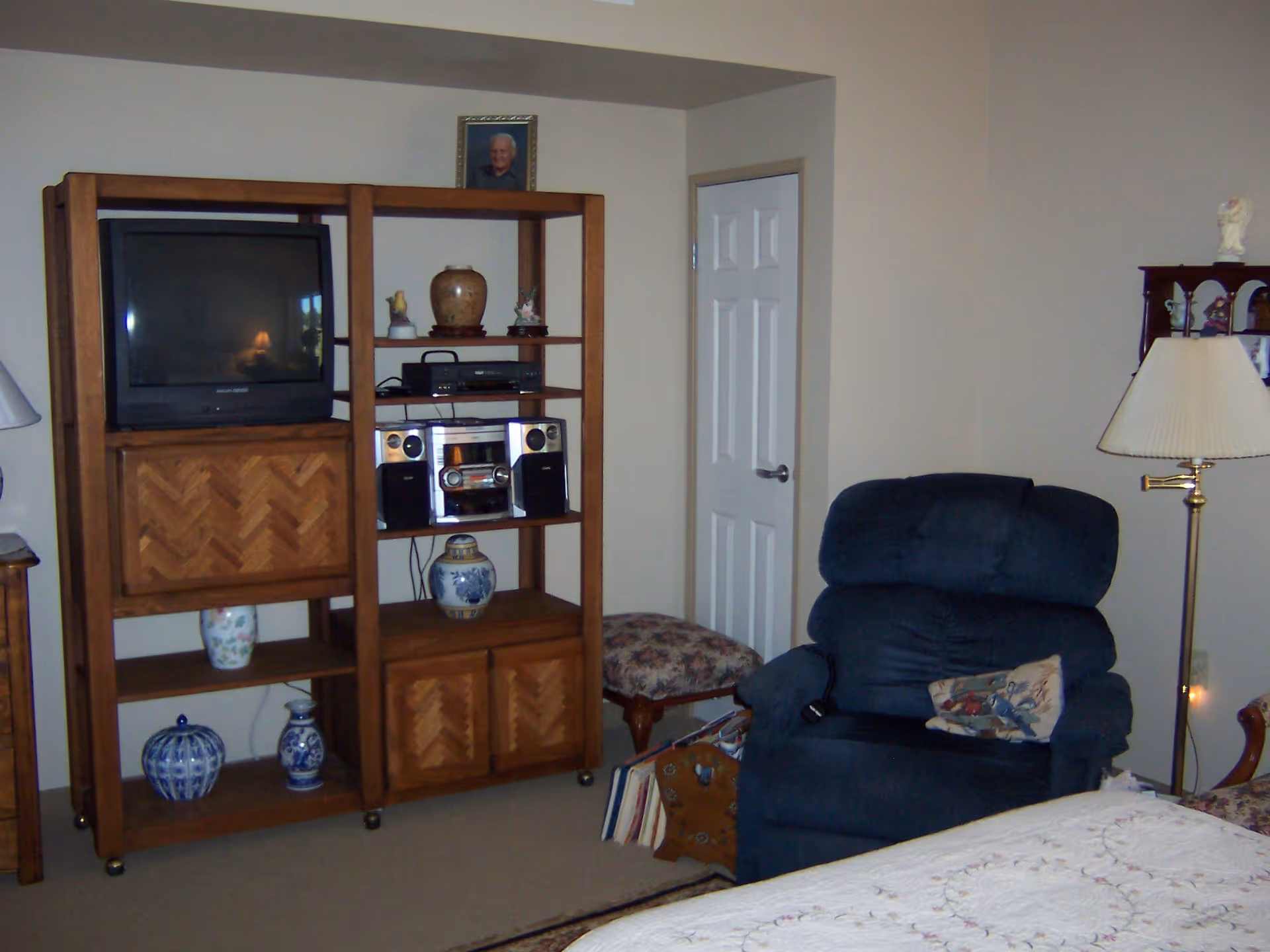 Room with wooden shelving holding a TV and stereo, a blue recliner, floor lamp, and part of a bed.