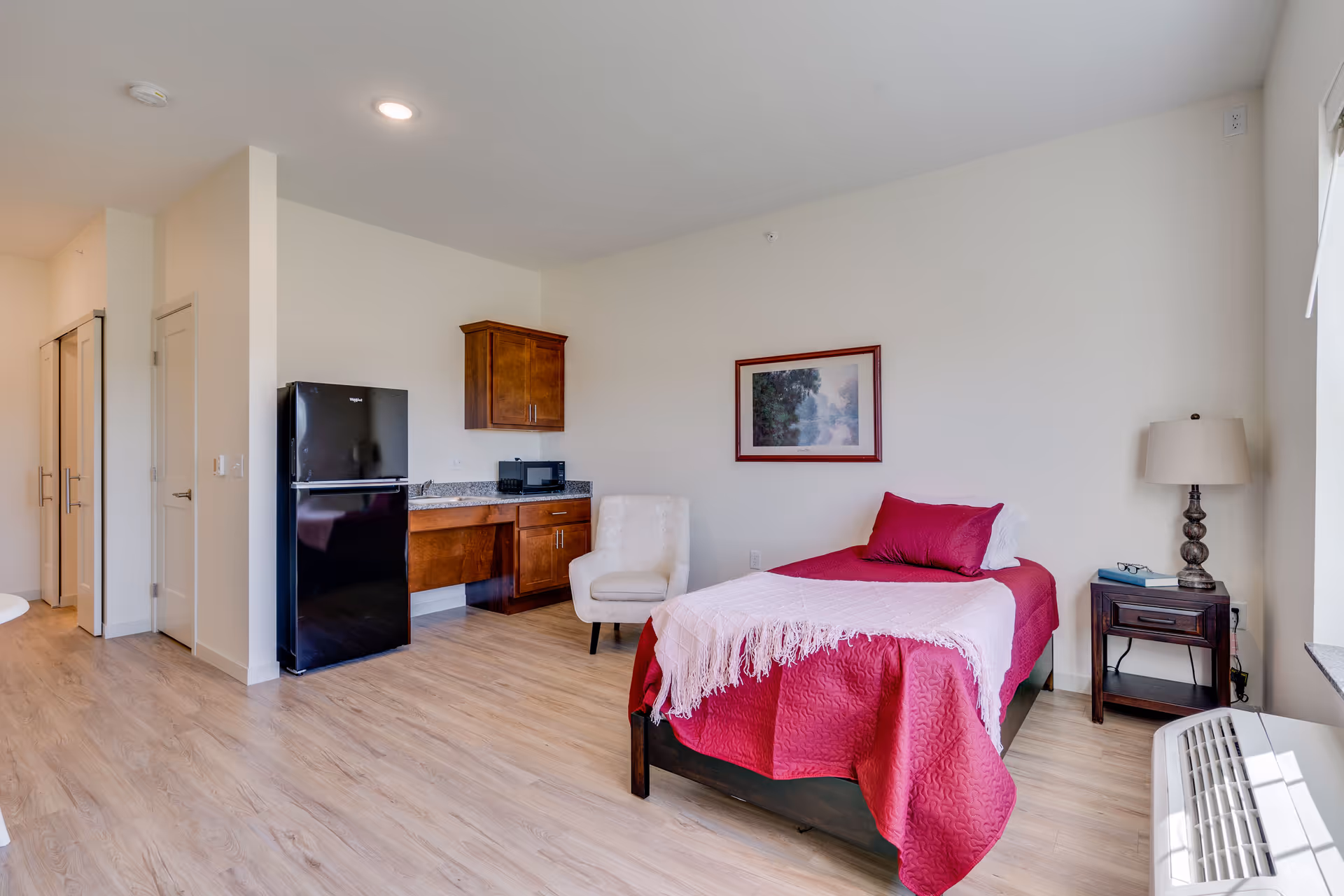 A senior living facility room with a single bed covered in a red bedspread and a light pink throw blanket. Next to the bed is a wooden nightstand with a lamp, a book, and a pair of glasses. There is a beige upholstered chair near a small kitchenette area with a black refrigerator, microwave, and wooden cabinets. The room has light wood flooring and white walls with a framed landscape painting above the bed.