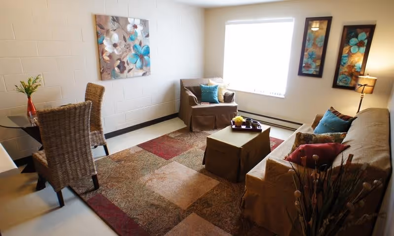 A cozy living room with a beige sofa and armchair, both adorned with colorful throw pillows. A rectangular ottoman serves as a coffee table with a tray on top. There is a patterned area rug covering the floor. A small dining table with two wicker chairs is placed near the wall, which has a floral painting. Two framed floral artworks and a floor lamp are positioned near the window, allowing natural light to brighten the room.