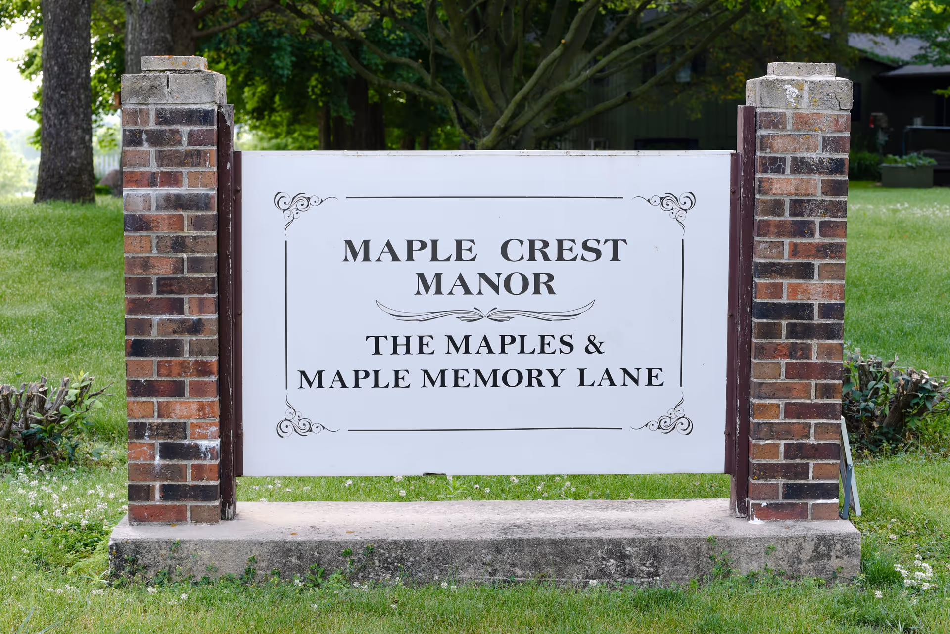 A white sign with decorative corners mounted between two brick pillars on a grassy area. The sign reads 'MAPLE CREST MANOR THE MAPLES & MAPLE MEMORY LANE' with trees and a building partially visible in the background.
