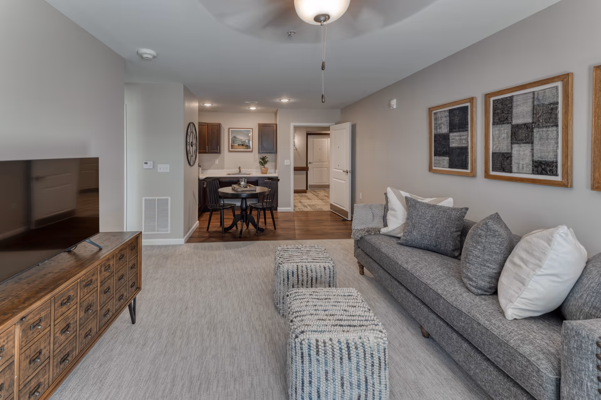 A modern senior living apartment interior featuring a gray sofa with multiple cushions, two woven ottomans, a wooden TV stand with a flat-screen TV, and a small dining area with a round table and two chairs. The walls are decorated with framed abstract artwork, and the flooring transitions from carpet in the living area to wood in the dining and kitchen area. The entrance door is open, showing a hallway beyond.
