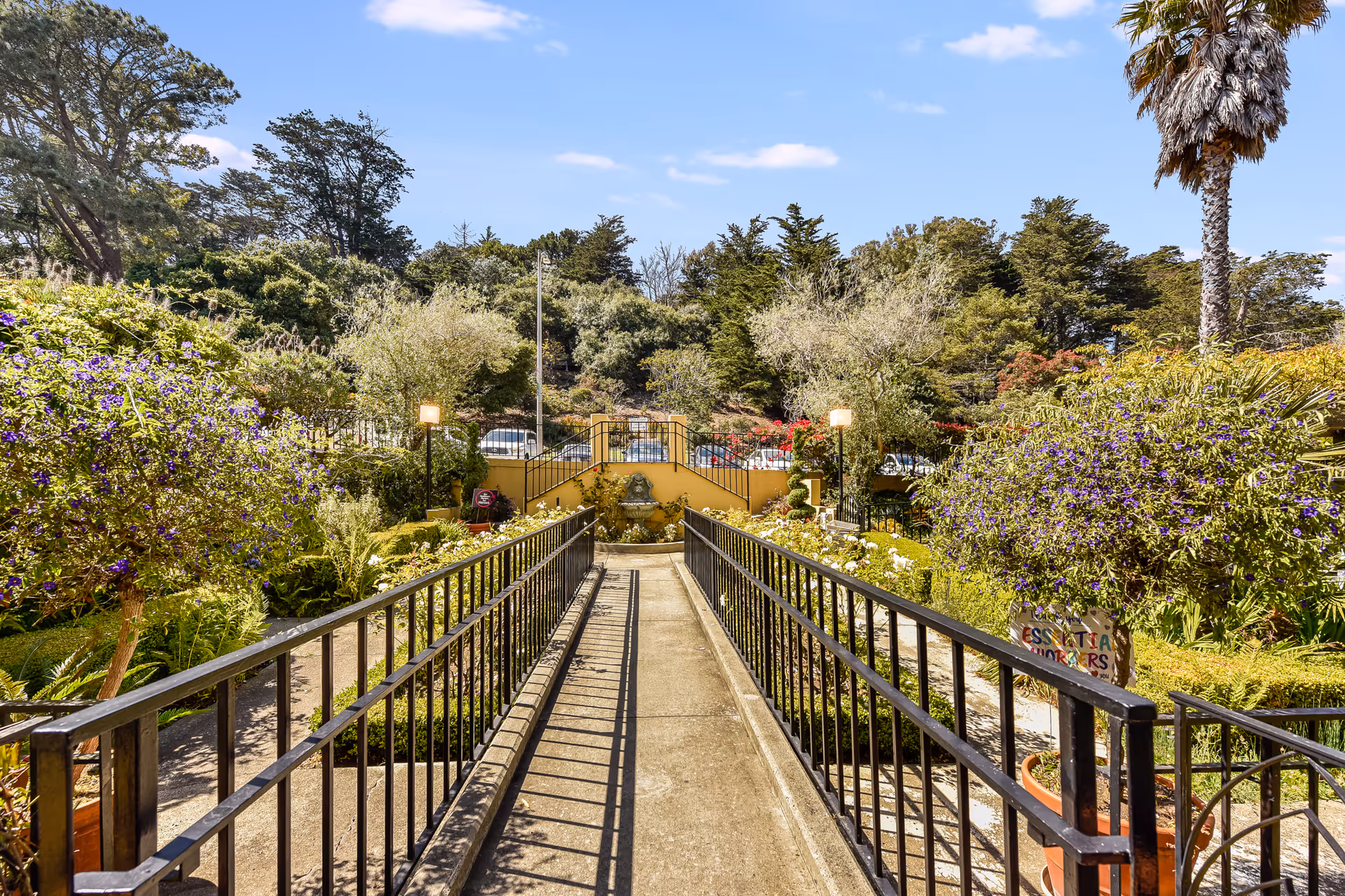 A sunny outdoor garden area with a concrete walkway bordered by black metal railings leading to a small yellow wall with a decorative fountain. The garden is lush with various green plants, purple flowers, and trees, including a tall palm tree on the right. The sky is clear with a few clouds.