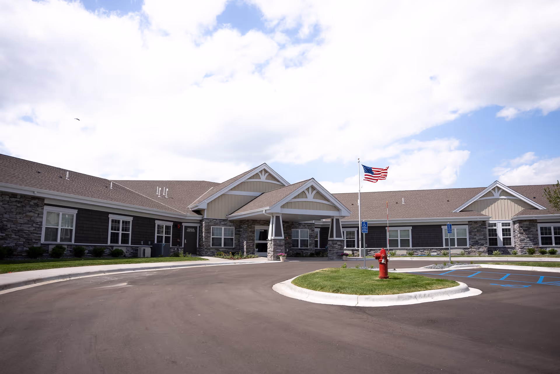 Exterior view of a single-story senior living facility building with stone and siding facade, a covered entrance, an American flag on a flagpole, a fire hydrant, and a parking area with handicap spaces under a partly cloudy sky.