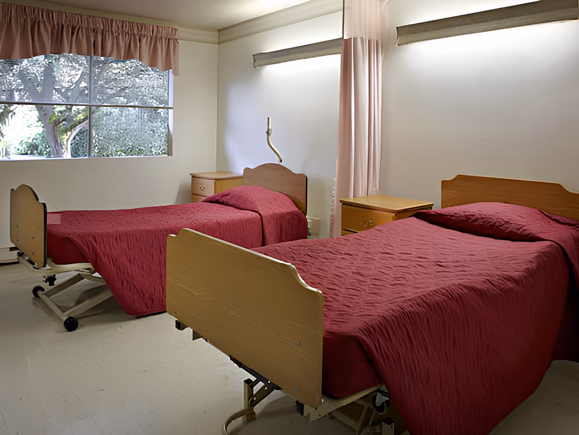 A room with two hospital-style beds covered in red bedspreads, each with a wooden headboard and footboard. There are two wooden nightstands between and beside the beds. A window with a pink valance and a curtain is on the left wall, and the walls are painted white.