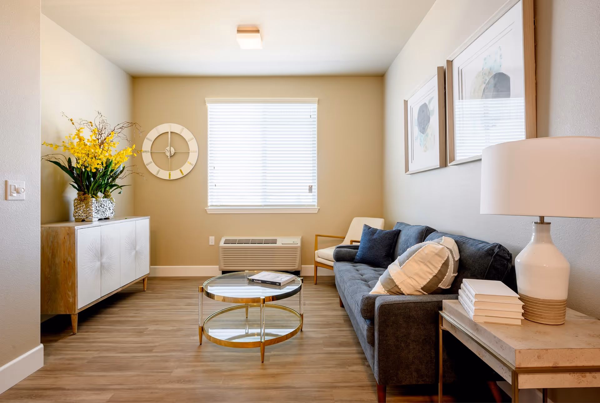 A cozy living room with a dark gray sofa adorned with multiple pillows, a glass coffee table with gold accents, a wooden side table with a white lamp and stacked books, and a wooden cabinet with a large vase of yellow flowers. The room has light beige walls, a window with white blinds, and two framed abstract artworks on the wall above the sofa.