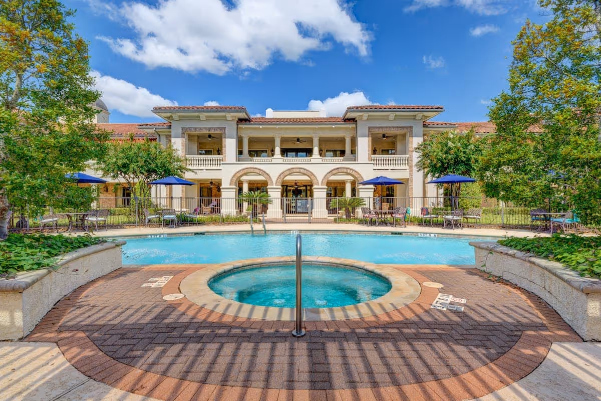 Outdoor swimming pool and hot tub area with lounge chairs and blue umbrellas, surrounded by greenery and trees, in front of a two-story building with arches and balconies under a partly cloudy blue sky.