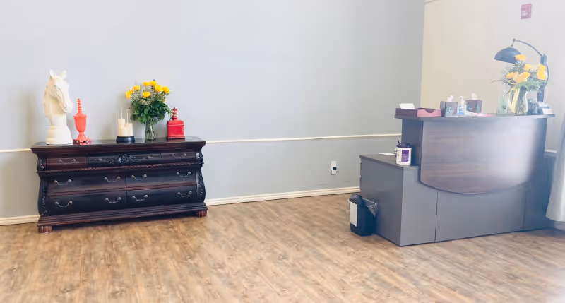 Reception area with wood-look flooring, a dark wooden dresser topped with flowers and decor on the left, and a curved reception desk with a lamp and flowers on the right.