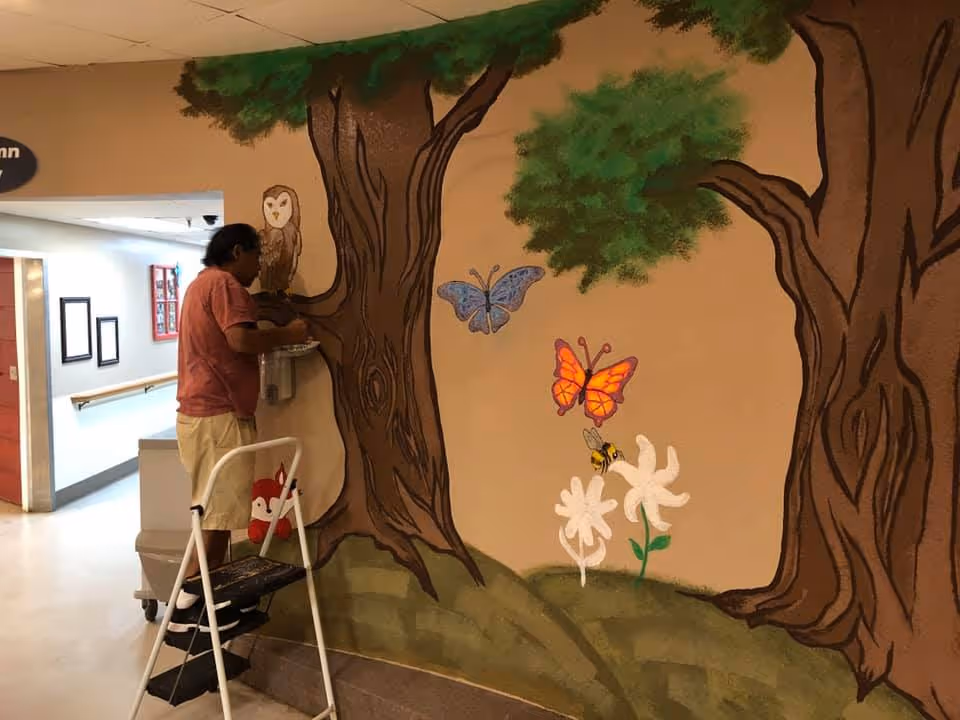 Person on a step stool painting a mural of trees, butterflies, and flowers on an indoor hallway wall.