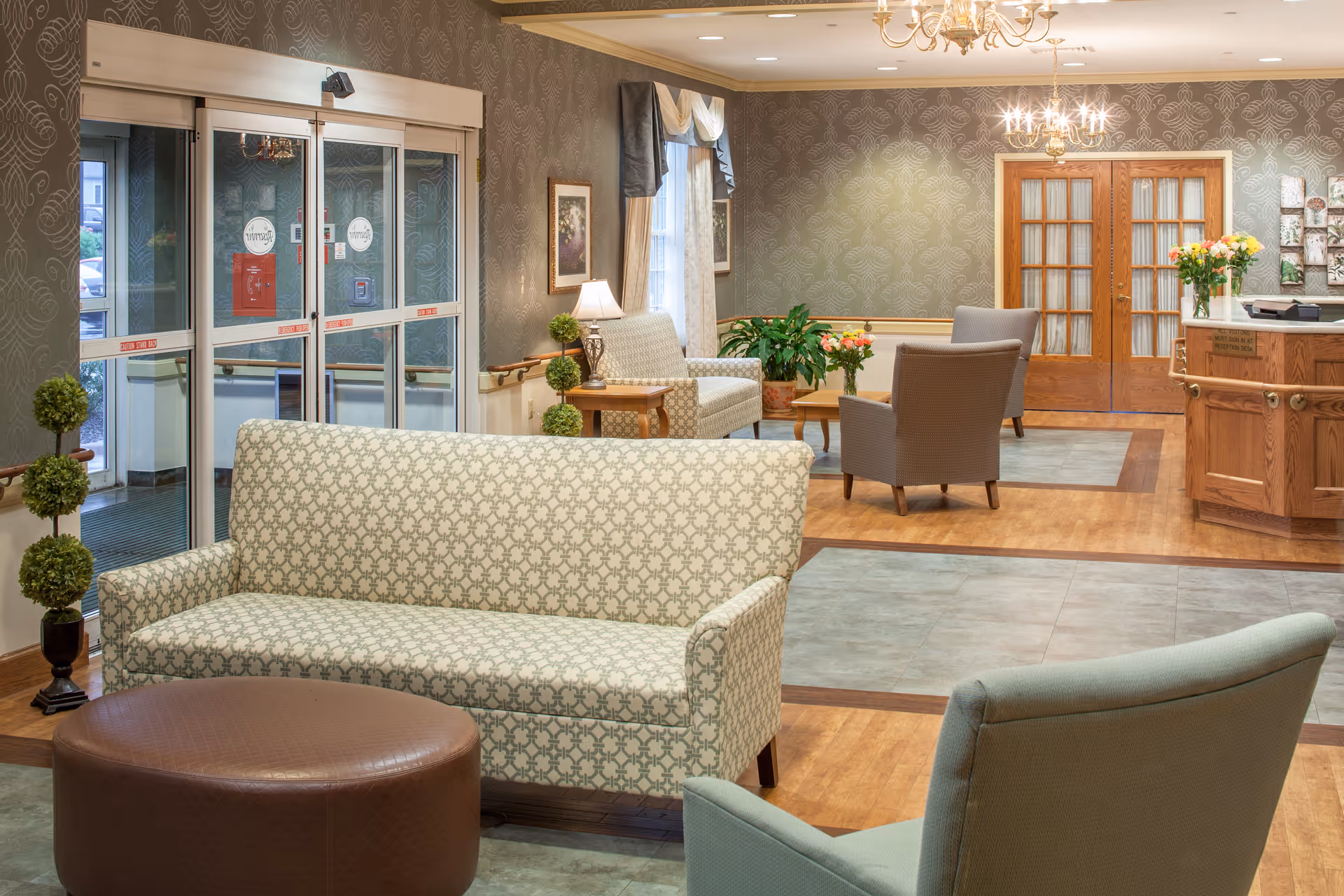 A well-lit and welcoming senior living facility lobby with patterned wallpaper, multiple upholstered chairs and sofas, a round brown ottoman, potted plants, a wooden reception desk with flowers, and glass double doors leading outside.