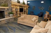 A cozy living room with two beige sofas facing each other on a patterned carpet. There is a stone fireplace on the left with decorative items on the mantel. A wooden built-in shelving unit with a television and various decor pieces is against the back wall. Two dark armchairs are positioned near a blue accent wall with circular wall art. Pendant lights hang from the ceiling.