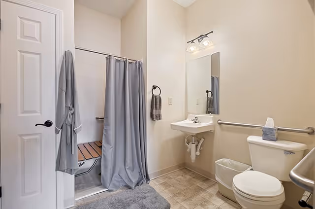 A clean and accessible bathroom featuring a toilet with a grab bar, a wall-mounted sink with a mirror above it, a shower area with a gray curtain and a wooden fold-down bench, a gray bathrobe hanging on the door, and a gray bath mat on the tiled floor.