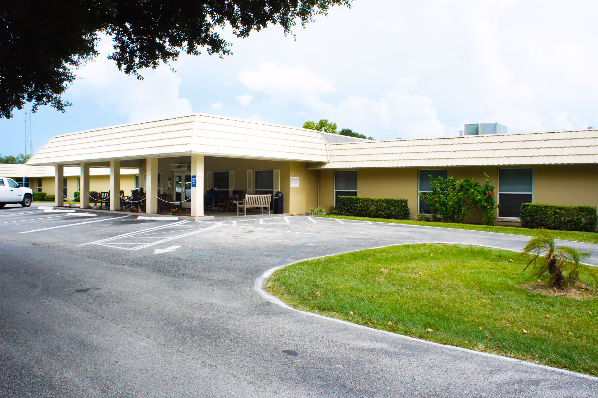 Front entrance of a single-story senior living building with a covered drive-up area, benches, parking spaces, and a grassy roundabout.
