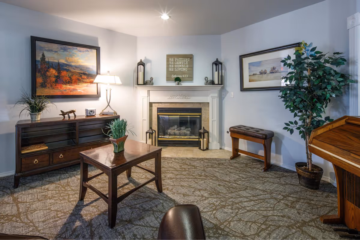 A cozy living room with a fireplace in the center, flanked by two black lanterns and decorative ducks. Above the fireplace is a wooden sign with an inspirational quote. To the left is a dark wooden cabinet with a table lamp, a small plant, and decorative items. A wooden coffee table with a potted plant sits in the middle of the carpeted floor. On the right side, there is a padded bench, a large potted plant, and a wooden piano or desk. The walls are light-colored and adorned with framed landscape paintings.