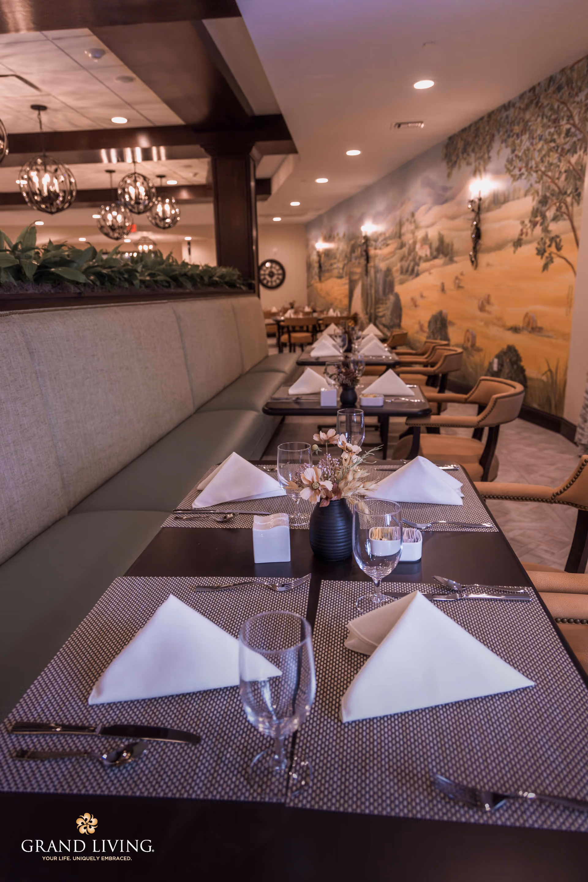 A neatly arranged dining area with tables set with white folded napkins, glassware, cutlery, and small floral centerpieces. The seating includes cushioned bench seating on one side and chairs on the other. The wall features a large mural with a nature scene, and decorative hanging lights illuminate the space.