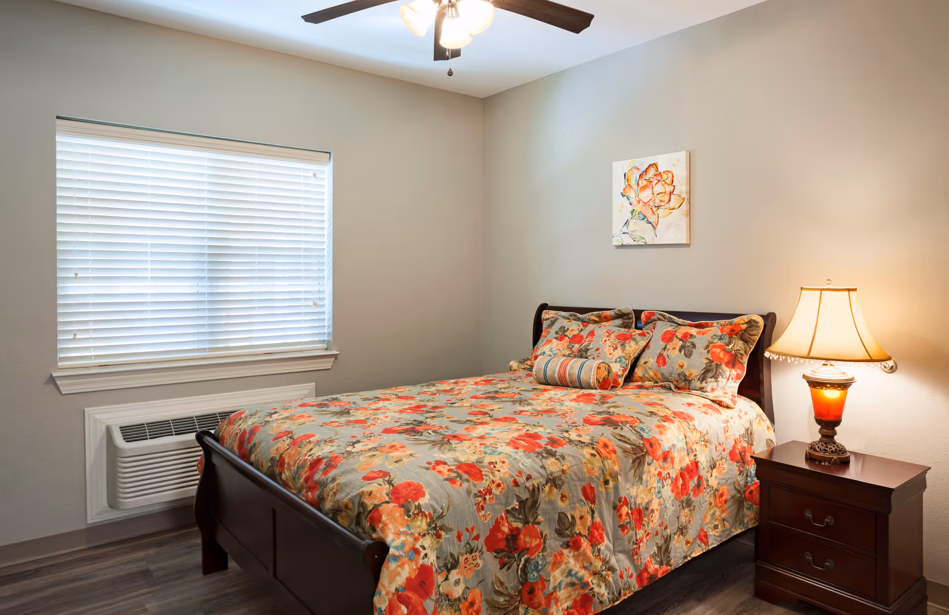 A cozy bedroom with a wooden bed frame and a floral-patterned bedspread in shades of red, orange, and green. There are matching pillows and a cylindrical bolster pillow on the bed. A wooden nightstand with two drawers is beside the bed, holding a decorative lamp with a warm light. A window with white blinds is on the left wall, and a ceiling fan with lights is mounted on the ceiling. A colorful floral painting hangs on the wall above the bed.