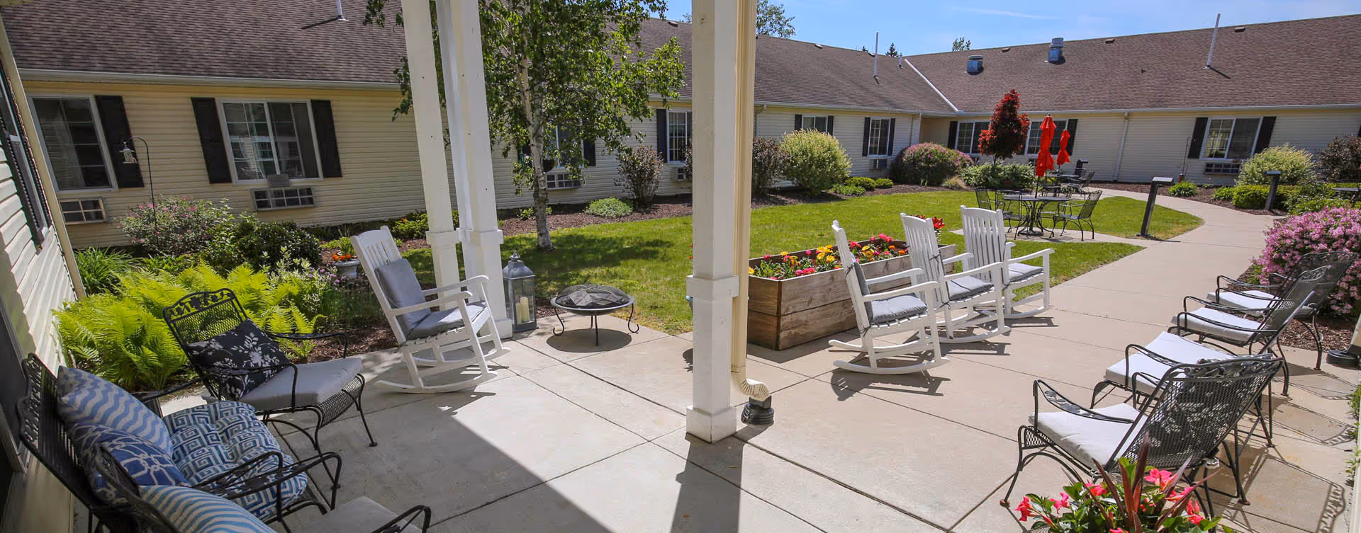 Outdoor patio area at Bickford of Saginaw Township with multiple chairs including rocking chairs and metal chairs with cushions arranged around a concrete walkway. There are flower beds, green grass, and shrubs surrounding the patio, with a building visible in the background under a clear blue sky.