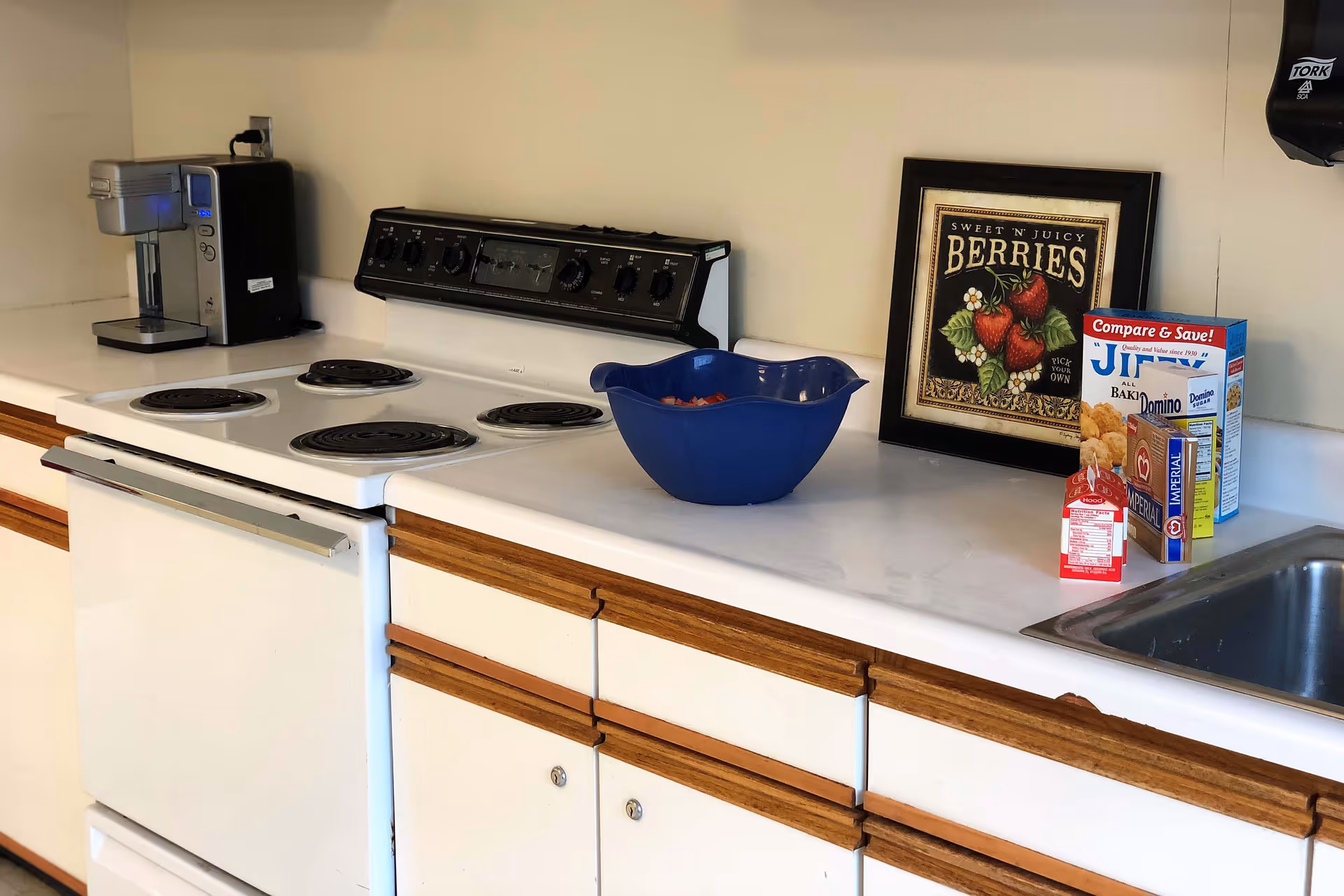 A kitchen countertop with a white electric stove, a coffee maker, a blue bowl, a framed picture of berries, various food items including a box of Jiffy corn muffin mix, Domino sugar, and a carton of milk, and a stainless steel sink. The cabinets below the countertop are white with wooden handles.