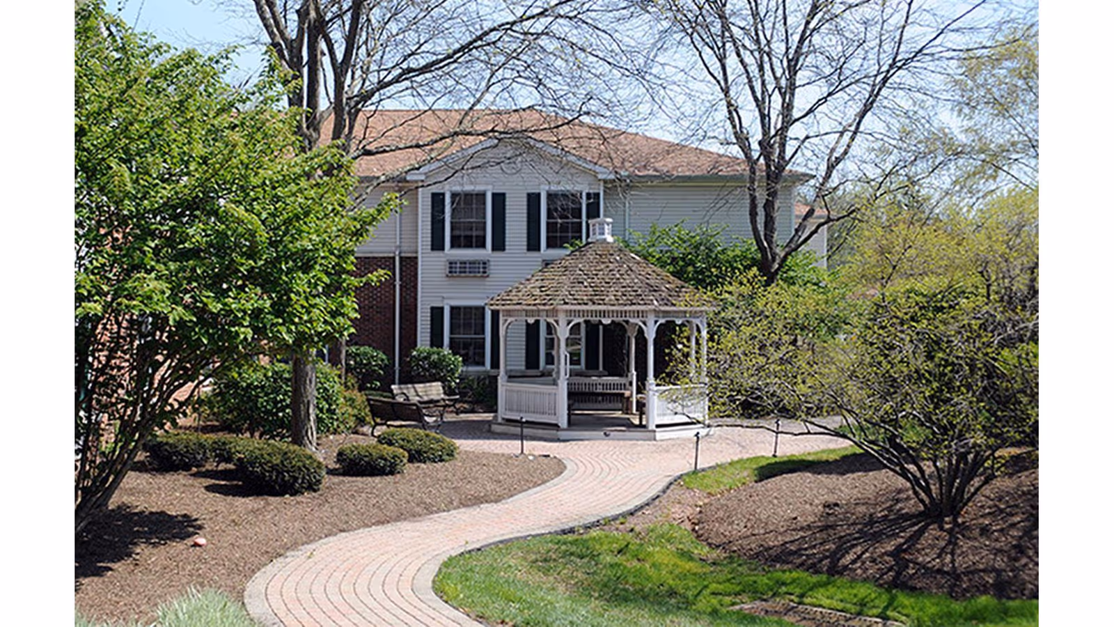 A winding brick pathway leads to a white gazebo with a shingled roof in a landscaped garden area with trees and bushes. In the background, there is a two-story building with white siding and black shutters.