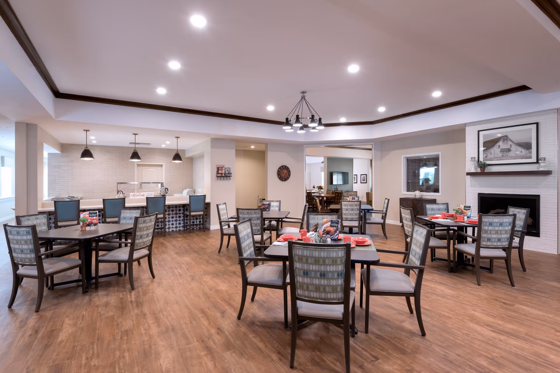 A spacious dining room with several tables and chairs arranged neatly. The tables are set with red plates and cups. The room features wooden flooring, a modern chandelier, recessed ceiling lights, and a fireplace with a framed picture above it. In the background, there is a counter with bar stools and pendant lights hanging above it.
