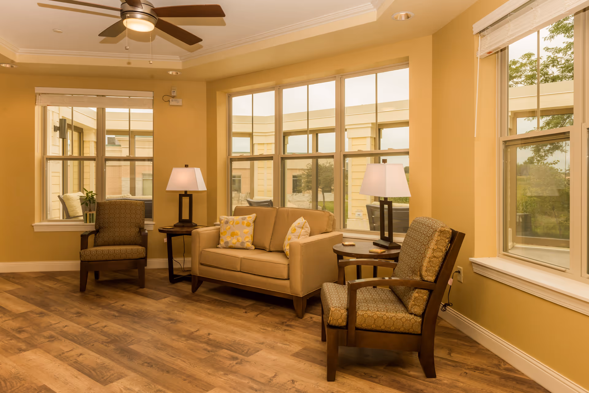 Sunlit communal sitting area with a sofa, two armchairs, side tables and lamps in front of large windows.